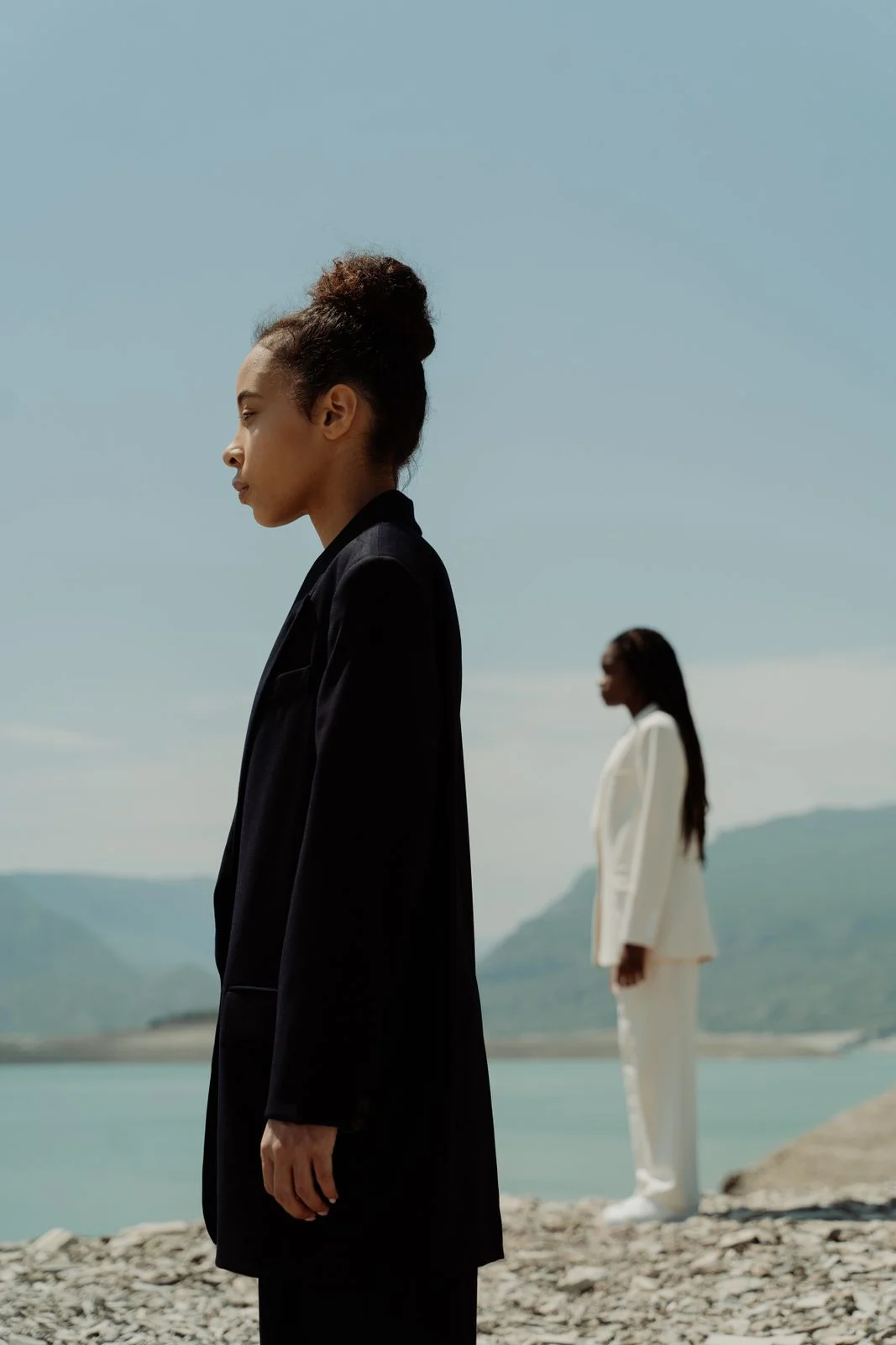 A model in a dark tailored jacket, with an ivory suit in the distance beside pale blue water.
